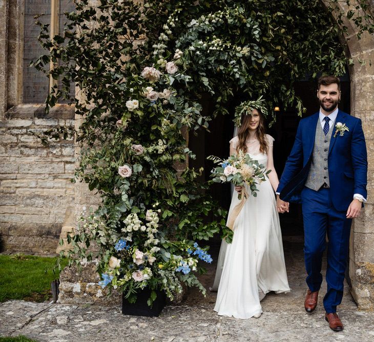 Bride in Charlie Brear Wedding Dress and Groom in Navy Ted Baker Suit Exiting the Church Decorated with Flowers