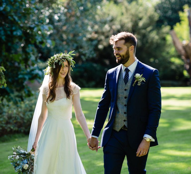 Bride in Charlie Brear Wedding Dress and Olive Flower Crown with Groom in Ted Baker Navy Suit Holding Hands