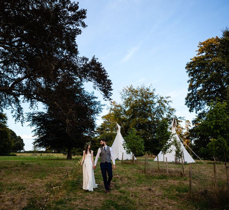 Bride in Charlie Brear Wedding Dress and Olive Flower Crown with Groom in Ted Baker Navy Suit Holding Hands Walking Through Glamping Field