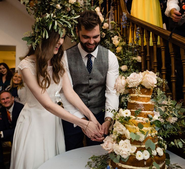 Bride in Charlie Brear Wedding Dress and Olive Flower Crown with Groom in Ted Baker Navy Suit Cutting the Naked Wedding Cake