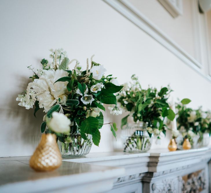 Fireplace Mantel Filled with Gold and Glass Vases filled with Green and White Flowers