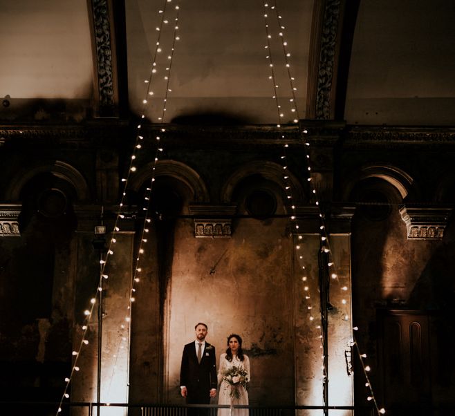 Bride in Grace Loves Lace Wedding Dress and Groom in Dark Suit Posing in Wilton's Music Hall with Fairy Lights