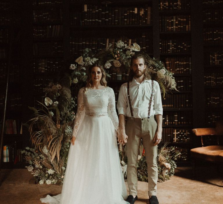 Bride in Separates and Groom in Chinos and Braces Standing in Front of a Dried Flower Moon Gate