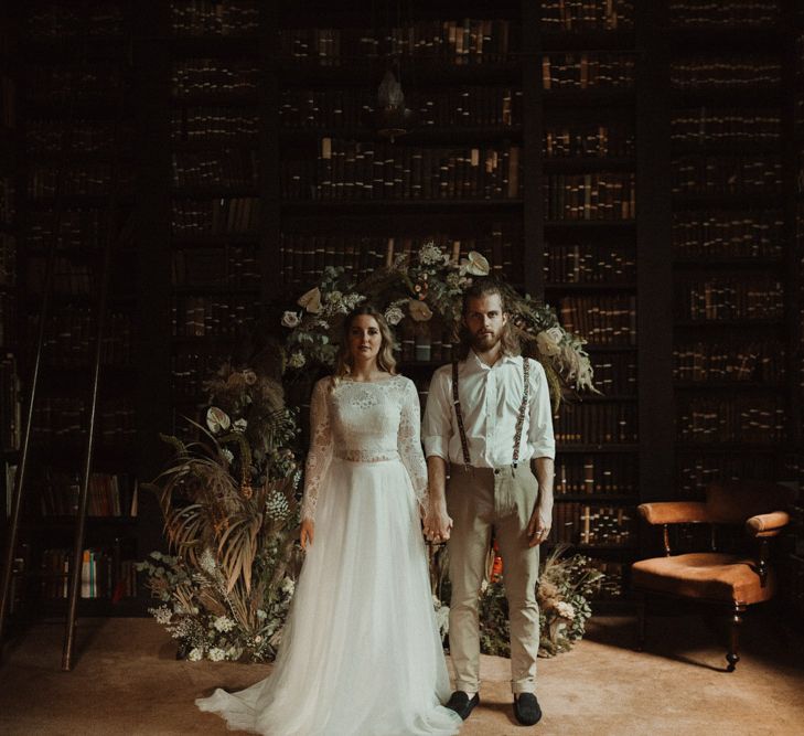 Bride in Separates and Groom in Chinos and Braces Standing in Front of a Dried Flower Moon Gate Holding Hands