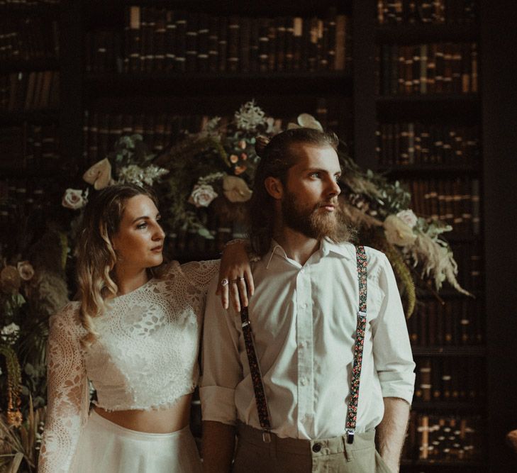 Bride in Separates and Groom in Chinos and Braces Standing in Front of a Dried Flower Moon Gate Embracing