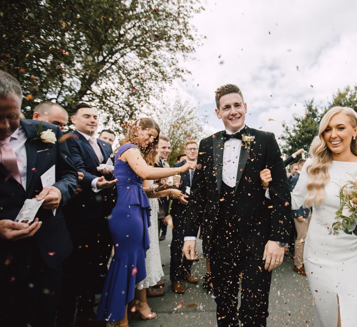 Bride and groom confetti exit with bride wearing split front dress and a long veil teamed with rose floral bouquet