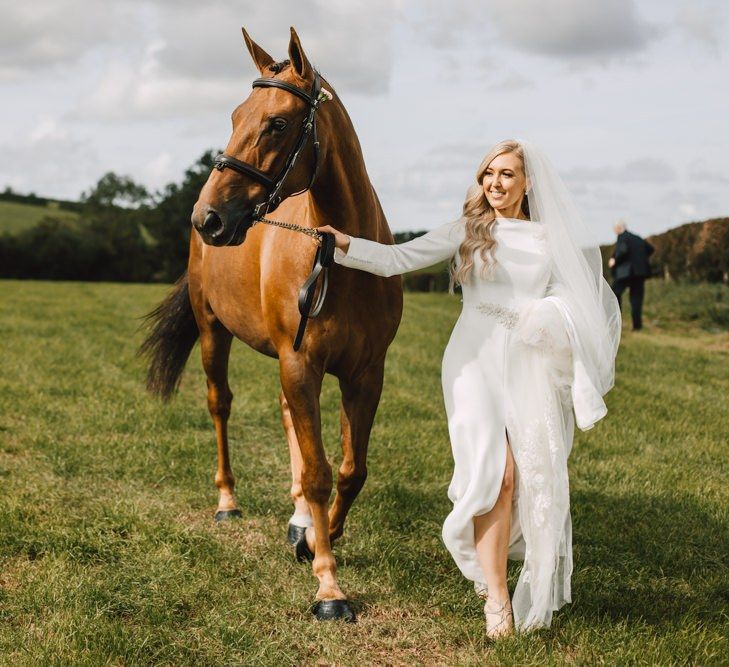 Bride wearing long lace tipped veil and split front bridal dress with her horse at marquee celebration with equestrian touches