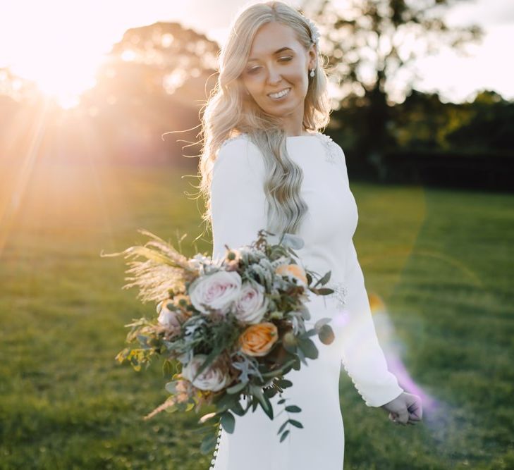 Bride wearing a simple split front wedding dress and peachy pink floral bouquet at marquee reception in autumn.