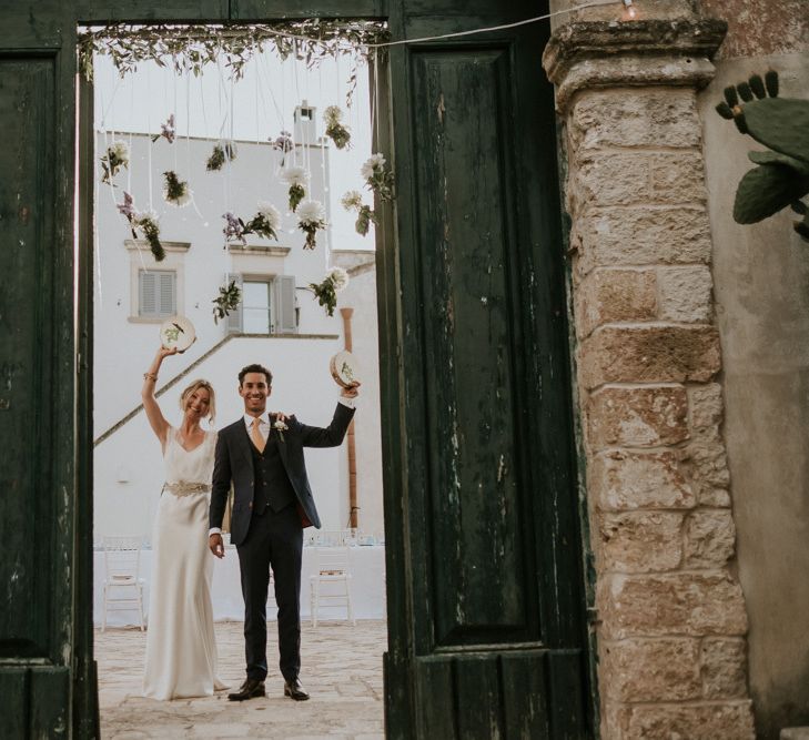 Bride in Halfpenny Wedding Dress and Groom in Navy Suit holding Tambourine's as They Enter The Wedding Breakfast