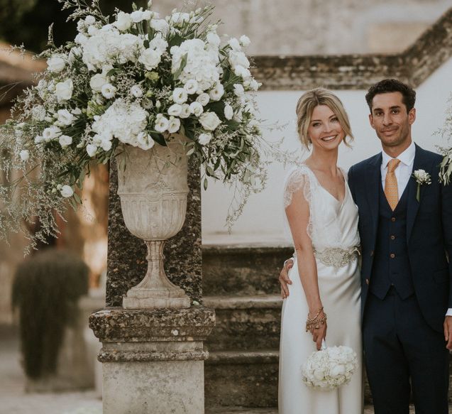 Bride in Halfpenny Wedding Dress and Groom in Navy Suit Posing By  All White Floral Arrangements