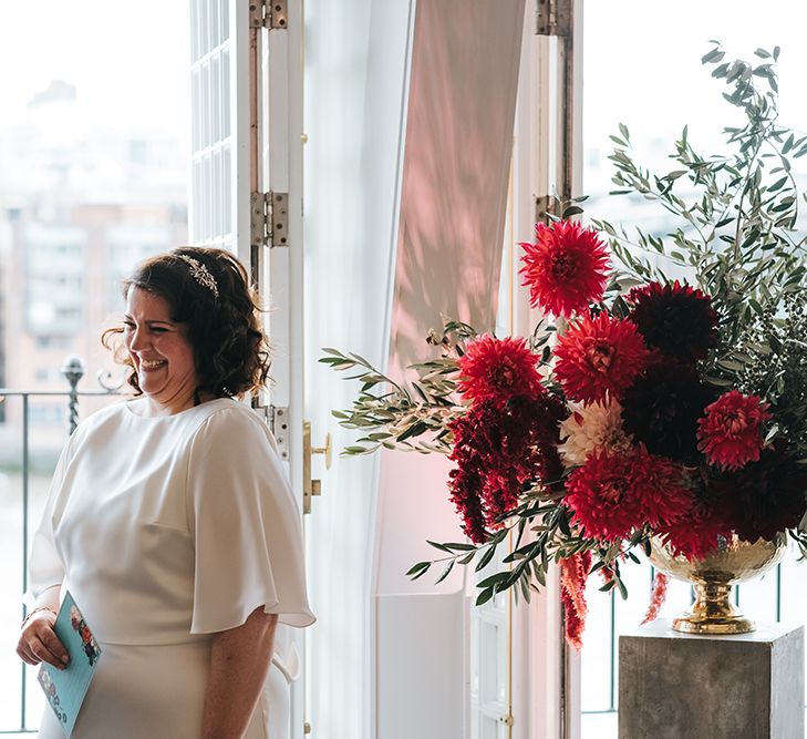 Bride in Halfpenny London Dress with Cowl Back and Half Length Sleeves | Tilly Thomas Lux Headband | Giant Urn of Red Flowers and Foliage on Grey Plinth | Wedding Ceremony at The Swan at The Globe | Meringue Kisses &amp; Colourful Wedding Flowers &amp; Stationery for a London Wedding at The Globe | Miss Gen