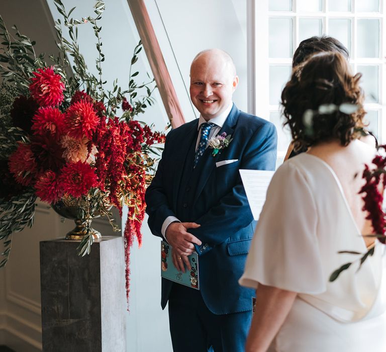 Bride in Halfpenny London Dress with Cowl Back and Half Length Sleeves | Groom in Blue Three-Piece Suit and White Trainers | Giant Urn of Red Flowers and Foliage on Grey Plinth | Wedding Ceremony at The Swan at The Globe | Meringue Kisses &amp; Colourful Wedding Flowers &amp; Stationery for a London Wedding at The Globe | Miss Gen