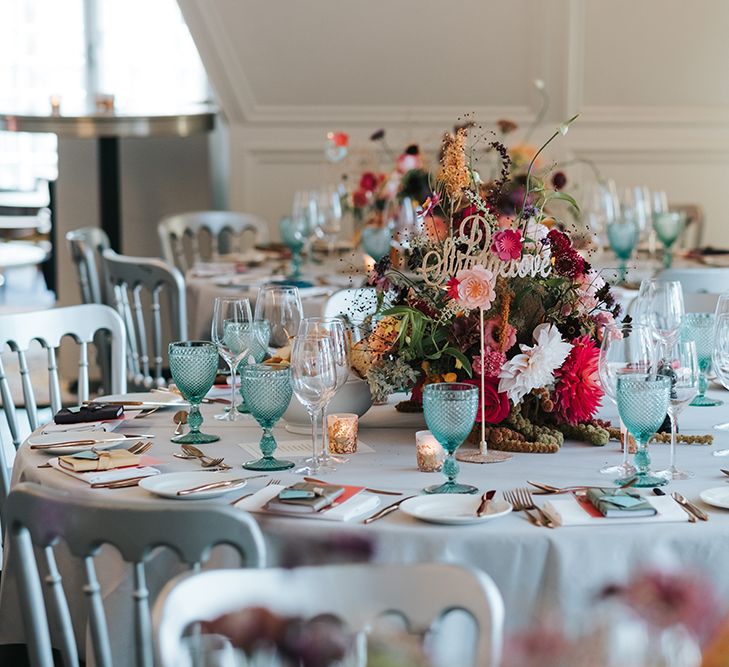 Sparkly Gold Table Sign | Colourful Floral Centrepiece | Tea Light Holders with Gold Glitter Detail | Blue Wine Glasses | Grey Tablecloth | Leather Book Wedding Favours | Personalised Pink Wedding Breakfast Menus | Duck Egg Blue Thank You Tags | Silver Chairs | Copper Cutlery | Meringue Kisses &amp; Colourful Wedding Flowers &amp; Stationery for a London Wedding at The Globe | Miss Gen