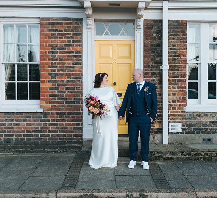 Bride in Halfpenny London Dress with Cowl Back and Half Length Sleeves | Colourful Deconstructed Bridal Bouquet of Red and Pink Flowers with Trailing Ribbon | Tilly Thomas Lux Headband | Groom in Blue Three-Piece Suit and White Trainers | Meringue Kisses &amp; Colourful Wedding Flowers &amp; Stationery for a London Wedding at The Globe | Miss Gen