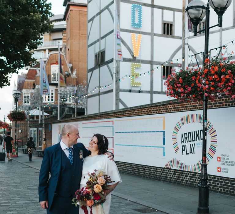 Bride in Halfpenny London Dress with Cowl Back and Half Length Sleeves | Colourful Deconstructed Bridal Bouquet of Red and Pink Flowers with Trailing Ribbon | Tilly Thomas Lux Headband | Groom in Blue Three-Piece Suit and White Trainers | Meringue Kisses &amp; Colourful Wedding Flowers &amp; Stationery for a London Wedding at The Globe | Miss Gen