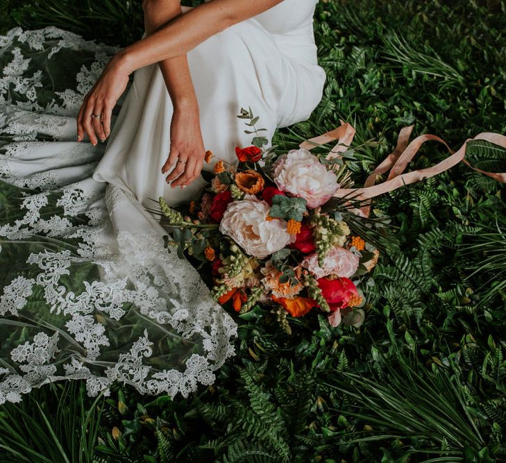 Bride Sitting on Foliage Next to Her Bright Bouquet Showing Off her Lace Wedding Dress Train