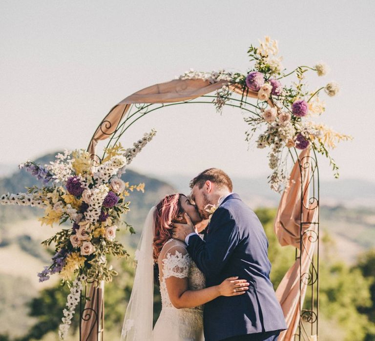 Bride and groom kiss at outdoor ceremony with flower arch