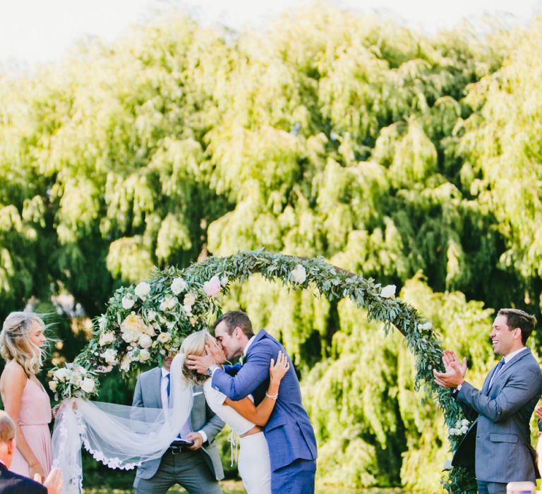 Bride in Fitted Noel and Jean Bridal Separates and Groom in Blue Suit Supply Suit Kissing in Front of Floral Moon Gate at Wedding Ceremony