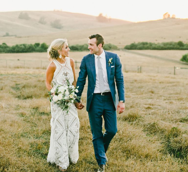 Bride in Lace Rue De Seine Wedding Dress and Groom in Blue Suit Walking Through the Fields