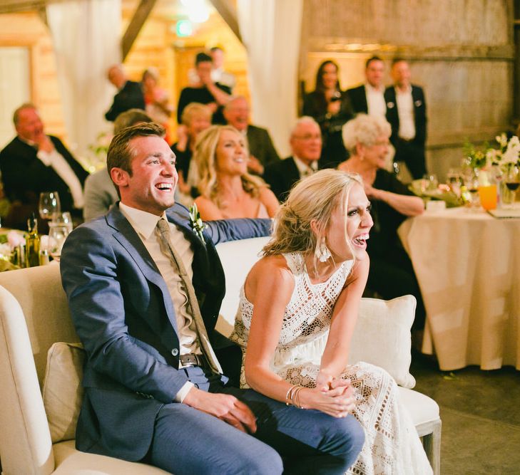 Bride in Lace Rue De Seine Wedding Dress and Groom in Blue Suit Laughing on Love Seat During Wedding Speeches