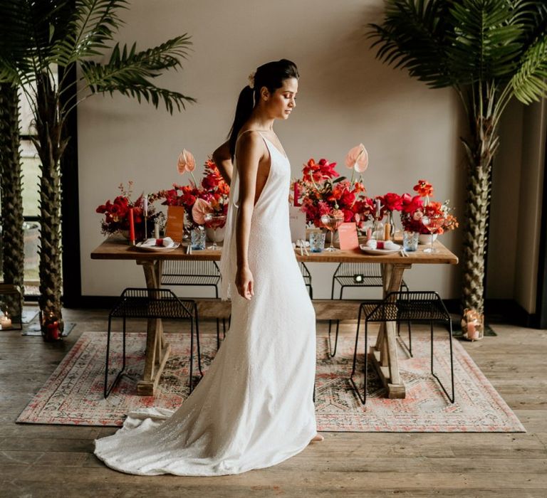 Bride in Minimalist Wedding Dress Standing in Front of a Tablescape filled with Red &amp; Pink Wedding Flowers