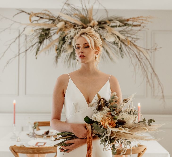 Bride in Minimalist Wedding Dress with Plunging Neckline Holding a Wedding Bouquet  of Dried Grasses, Foliage and Orange Flowers