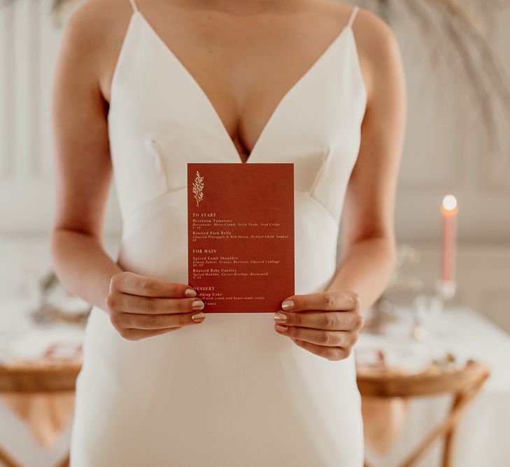 Bride in Minimalist Wedding Dress with Plunging Neckline Holding a Rust Coloured Wedding Menu Card