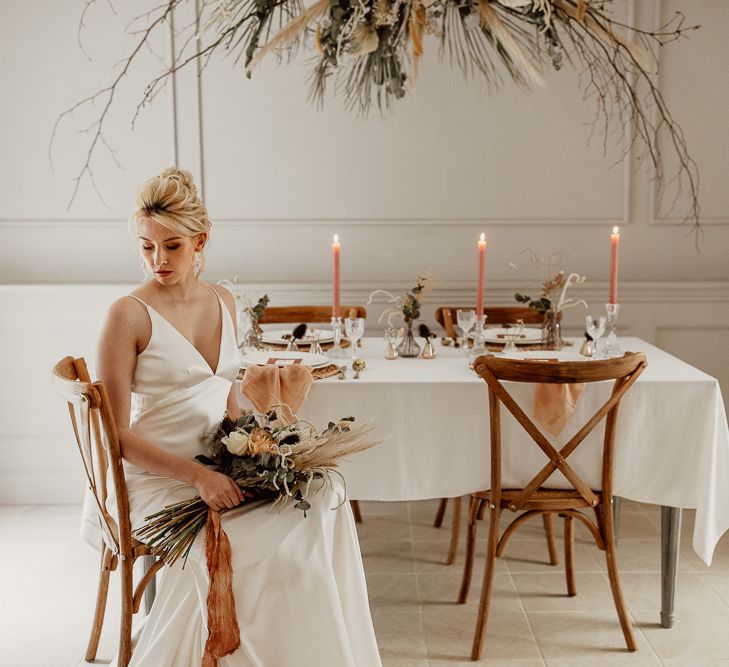 Bride in Minimalist Wedding Dress Sitting at a Tablescape with Flower Installation and Taper Candles