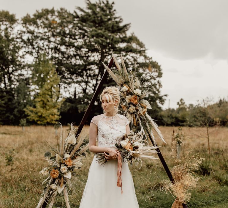 Bride in Tulle Skirt and Lace Top Wedding Dress Standing in From of a Wooden Frame Altar Decorated in Flowers