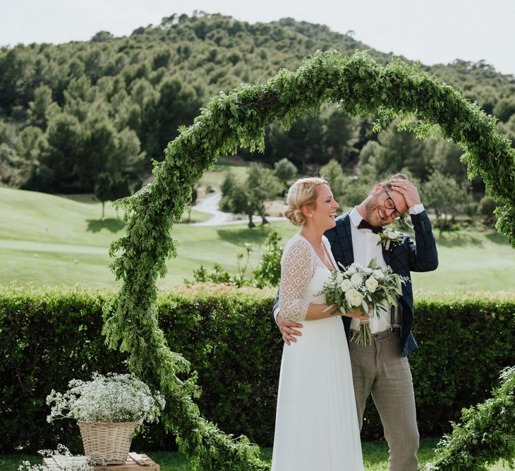 Bride in Rembo Styling Wedding Dress  and Groom  in Chino's and Checked Blazer Standing in Front of Foliage Covered Moon Gate