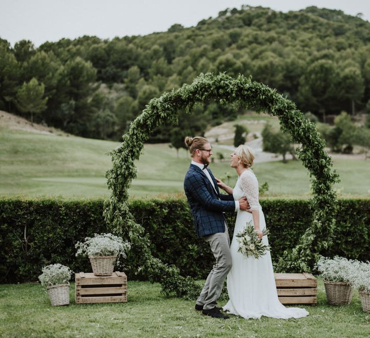 Bride in Rembo Styling Wedding Dress and Groom  in Chino's and Checked Blazer Standing in Front of A Greenery Moon Gate