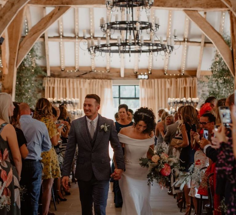 Bride in off the shoulder wedding dress and groom in blue suit walking up the aisle as husband and wife