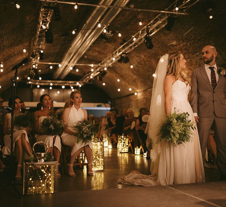 Wedding Ceremony | Bride in  Bespoke Wedding Gown with Blush Underskirt and One Off Shoulder Strap | Fingertip Length Veil Customised with Feathers | Nude Topshop Shoes with Pom Pom | Homemade Bouquet of White Flowers, Ferns and Foliage | Groom in Grey Puppy Tooth Check Two-Piece Paul Smith Suit with Amber Tie Pin, Pocket Square and Braces | Lanterns Filled with Fairylights | Festoon Lights | Bike Shed Motorcycle Club Wedding for ELLE Digital Editor | Nigel John Photography