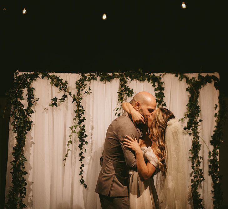 First Kiss | Altar with White Drapery and Foliage Garland | Bride in  Bespoke Wedding Gown with Blush Underskirt and One Off Shoulder Strap | Fingertip Length Veil Customised with Feathers | Groom in Grey Puppy Tooth Check Two-Piece Paul Smith Suit with Amber Tie Pin, Pocket Square and Braces | Bike Shed Motorcycle Club Wedding for ELLE Digital Editor | Nigel John Photography