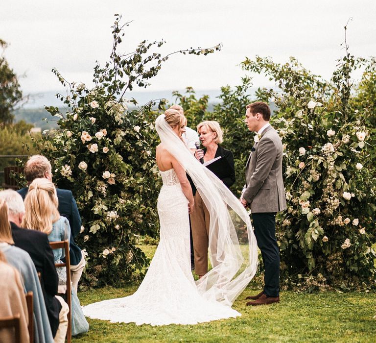 Bride and groom at the alter with white floral arch and long veil