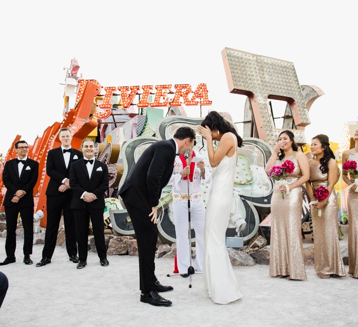 Bride in Silk BHLDN Dress with Spaghetti Straps and V-Back | Groom in Black Tuxedo with Emerald Green Bow Tie | Bridesmaids in Gold Sequin Maxi Dresses with Cowl Back | Fuschia Sweet Pea Spray Silk Flower Bouquets |Groomsmen in Black Tuxedos | Elvis Presley Celebrant | Wedding Ceremony at The Neon Museum | Las Vegas Wedding with Gold Sequin Bridesmaids Dresses and Silk Flowers | Chris Barber Photography