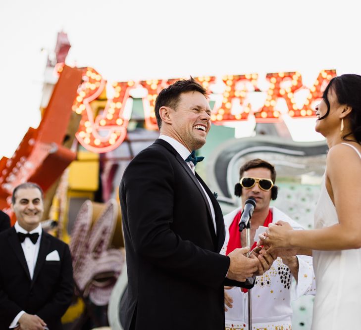 Bride in Silk BHLDN Dress with Spaghetti Straps and V-Back | Groom in Black Tuxedo with Emerald Green Bow Tie | Elvis Presley Celebrant | Groomsmen in Black Tuxedos | Wedding Ceremony at The Neon Museum | Las Vegas Wedding with Gold Sequin Bridesmaids Dresses and Silk Flowers | Chris Barber Photography