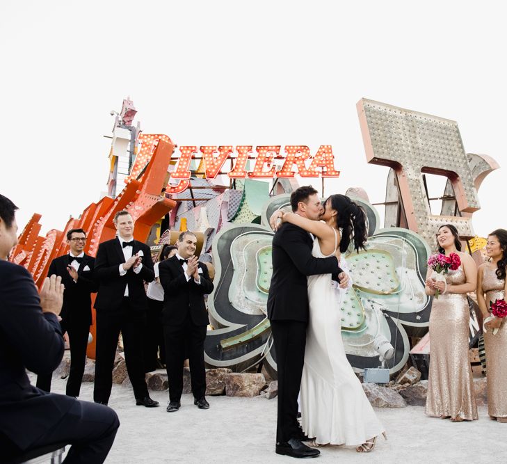 Bride in Silk BHLDN Dress with Spaghetti Straps and V-Back | Groom in Black Tuxedo with Emerald Green Bow Tie | Bridesmaids in Gold Sequin Maxi Dresses with Cowl Back | Fuschia Sweet Pea Spray Silk Flower Bouquets | Groomsmen in Black Tuxedos | Wedding Ceremony at The Neon Museum | Las Vegas Wedding with Gold Sequin Bridesmaids Dresses and Silk Flowers | Chris Barber Photography