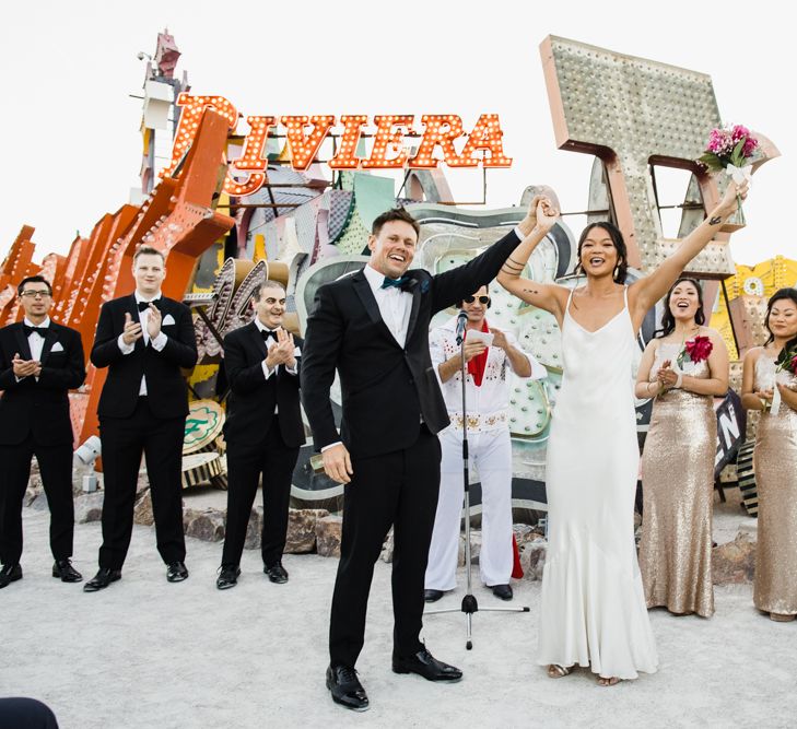 Bride in Silk BHLDN Dress with Spaghetti Straps and V-Back | Groom in Black Tuxedo with Emerald Green Bow Tie | Bridesmaids in Gold Sequin Maxi Dresses with Cowl Back | Fuschia Sweet Pea Spray Silk Flower Bouquets | Groomsmen in Black Tuxedos | Elvis Presley Celebrant | Wedding Ceremony at The Neon Museum | Las Vegas Wedding with Gold Sequin Bridesmaids Dresses and Silk Flowers | Chris Barber Photography