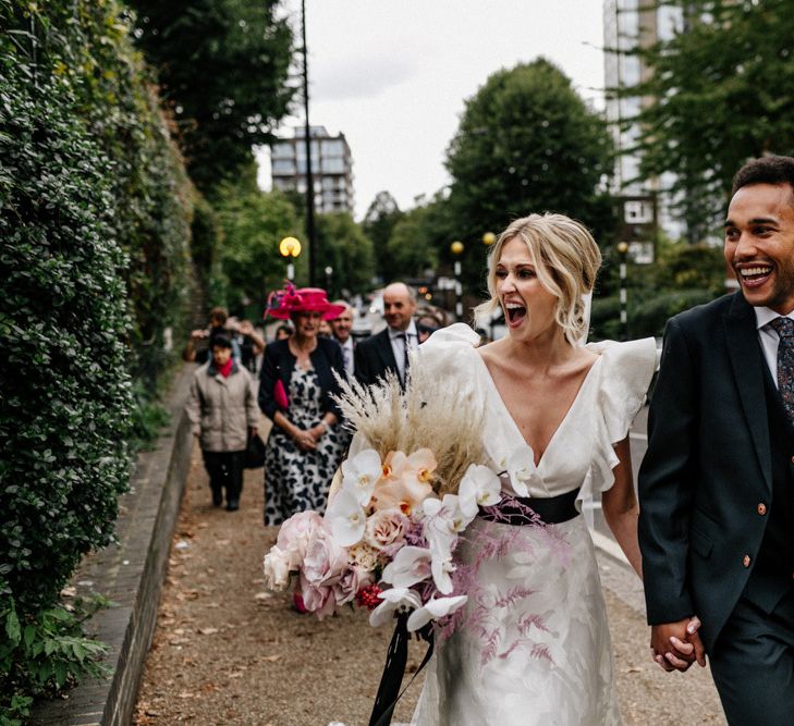 Stylish Bride and Groom Walking From Their Ceremony to Their Reception in Ruffle Wedding Dress and Navy Suit