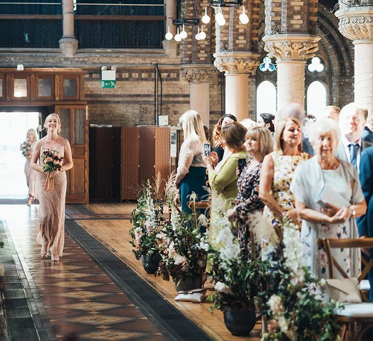 Bridesmaids wearing peach embellished dresses walking down the aisle at church ceremony with fresh foliage decor