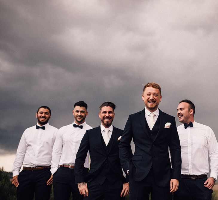 Groom and Best Man in Three-Piece Hawes &amp; Curtis Suit with Ivory Tie and Pocket Square | Groomsmen in Shirts and Bow Ties | Rue de Seine Wedding Dress with Pronovias Overskirt, Personalised Place Setting Illustrations and Sweetheart Table | Samuel Docker Photography