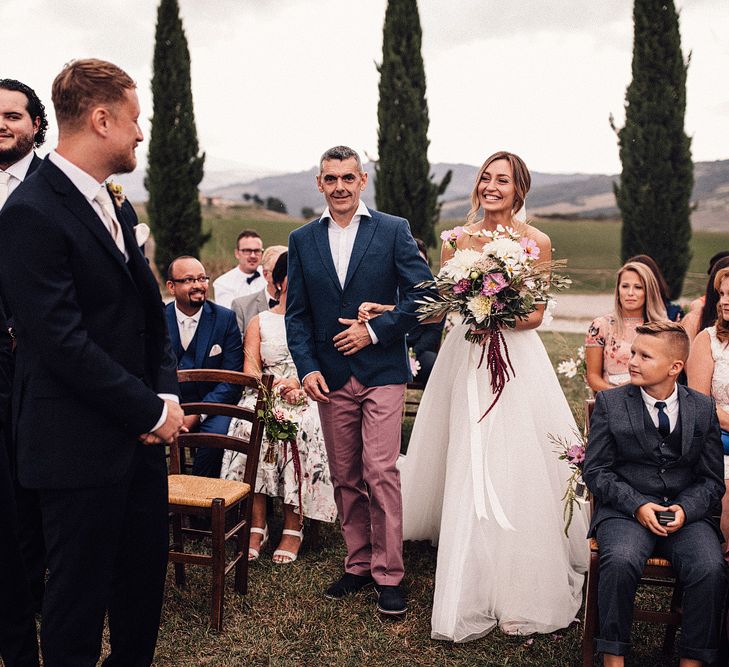 Bride in Structured Bustier Rue de Seine Fox Gown with Floral Appliqué Detail and Off The Shoulder Straps | Pronovias Chiffon Overskirt | Pink, Purple and White Wildflower Bouquet with White Trailing Ribbon | Groom in Three-Piece Hawes &amp; Curtis Suit with Ivory Tie and Pocket Square | Outdoor Wedding Ceremony at Il Rigo Agriturismo, Tuscany | Rue de Seine Wedding Dress with Pronovias Overskirt, Personalised Place Setting Illustrations and Sweetheart Table | Samuel Docker Photography