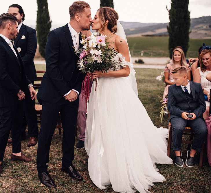 Bride in Structured Bustier Rue de Seine Fox Gown with Floral Appliqué Detail and Off The Shoulder Straps | Pronovias Chiffon Overskirt | Pink, Purple and White Wildflower Bouquet with White Trailing Ribbon | Groom in Three-Piece Hawes &amp; Curtis Suit with Ivory Tie and Pocket Square | Outdoor Wedding Ceremony at Il Rigo Agriturismo, Tuscany | Rue de Seine Wedding Dress with Pronovias Overskirt, Personalised Place Setting Illustrations and Sweetheart Table | Samuel Docker Photography