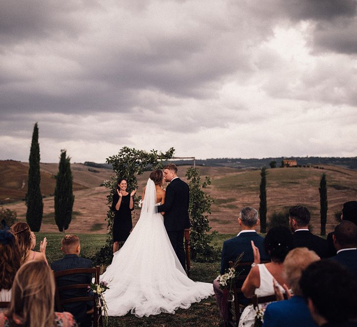 Bride in Structured Bustier Rue de Seine Fox Gown with Floral Appliqué Detail and Off The Shoulder Straps | Pronovias Chiffon Overskirt | Groom in Three-Piece Hawes &amp; Curtis Suit with Ivory Tie and Pocket Square | Wooden Wedding Arch with Botanicals | Outdoor Wedding Ceremony at Il Rigo Agriturismo, Tuscany | Rue de Seine Wedding Dress with Pronovias Overskirt, Personalised Place Setting Illustrations and Sweetheart Table | Samuel Docker Photography