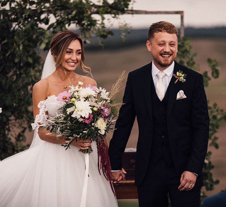 Bride in Structured Bustier Rue de Seine Fox Gown with Floral Appliqué Detail and Off The Shoulder Straps | Pronovias Chiffon Overskirt | Pink, Purple and White Wildflower Bouquet with White Trailing Ribbon | Groom in Three-Piece Hawes &amp; Curtis Suit with Ivory Tie and Pocket Square | Wooden Wedding Arch with Botanicals | Il Rigo Agriturismo, Tuscany | Rue de Seine Wedding Dress with Pronovias Overskirt, Personalised Place Setting Illustrations and Sweetheart Table | Samuel Docker Photography