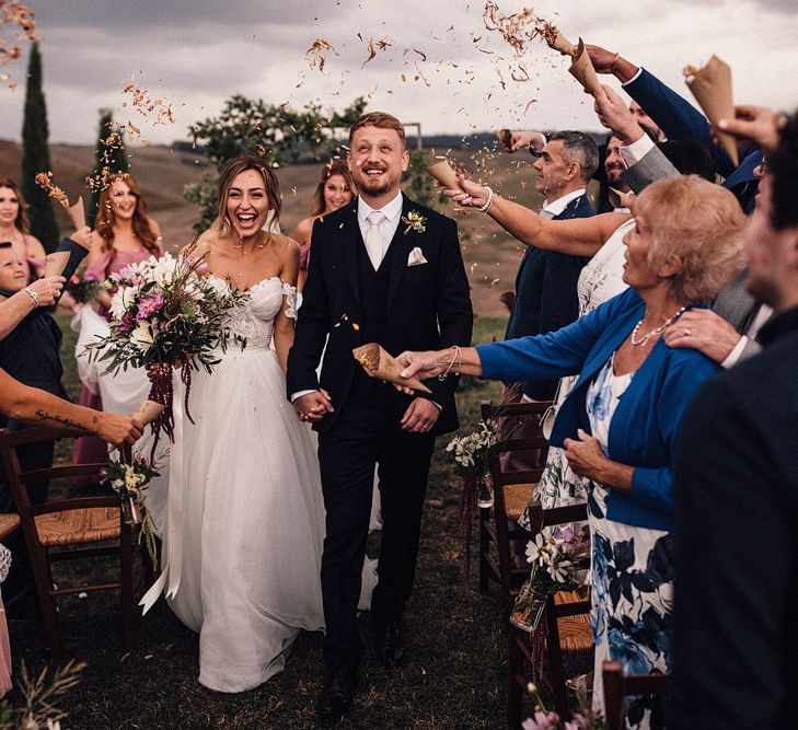 Bride in Structured Bustier Rue de Seine Fox Gown with Floral Appliqué Detail and Off The Shoulder Straps | Pronovias Chiffon Overskirt | Pink, Purple and White Wildflower Bouquet with White Trailing Ribbon | Groom in Three-Piece Hawes &amp; Curtis Suit with Ivory Tie and Pocket Square | Wooden Wedding Arch with Botanicals | Il Rigo Agriturismo, Tuscany | Rue de Seine Wedding Dress with Pronovias Overskirt, Personalised Place Setting Illustrations and Sweetheart Table | Samuel Docker Photography