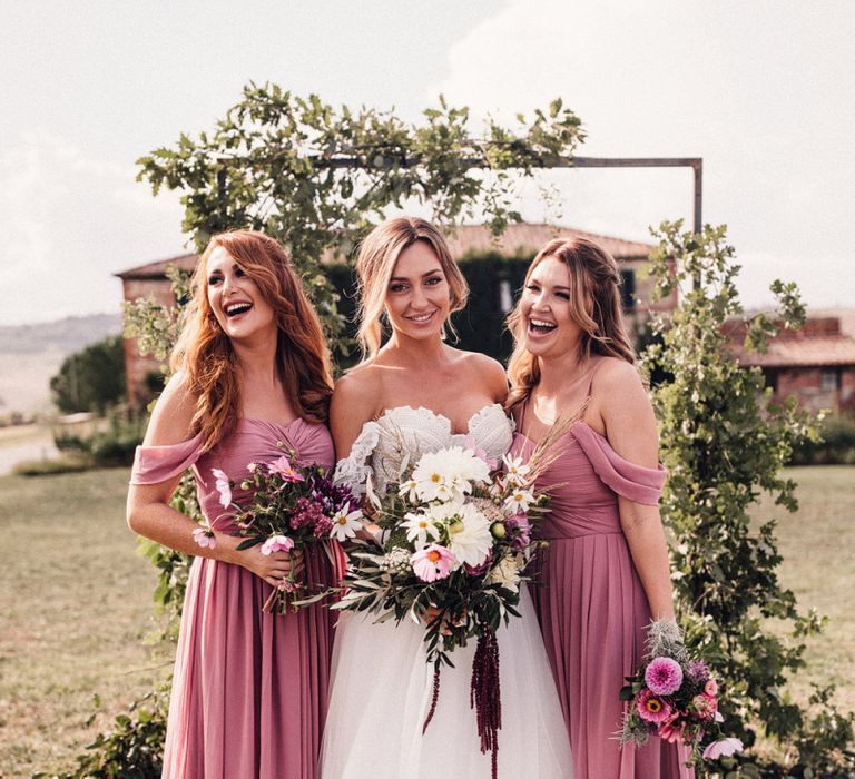 Bride in Structured Bustier Rue de Seine Fox Gown with Floral Appliqué Detail and Off The Shoulder Straps | Pronovias Chiffon Overskirt | Pink, Purple and White Wildflower Bouquet with White Trailing Ribbon | Bridesmaids in Dusky Pink ASOS Dresses with Ruched Bodice and Off-Shoulder Straps | Wooden Wedding Arch with Botanicals | Il Rigo Agriturismo | Rue de Seine Wedding Dress with Pronovias Overskirt, Personalised Place Setting Illustrations and Sweetheart Table | Samuel Docker Photography