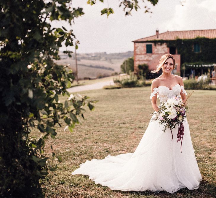 Bride in Structured Bustier Rue de Seine Fox Gown with Floral Appliqué Detail and Off The Shoulder Straps | Pronovias Chiffon Overskirt | Pink, Purple and White Wildflower Bouquet with White Trailing Ribbon | Il Rigo Agriturismo, Tuscany | Rue de Seine Wedding Dress with Pronovias Overskirt, Personalised Place Setting Illustrations and Sweetheart Table | Samuel Docker Photography