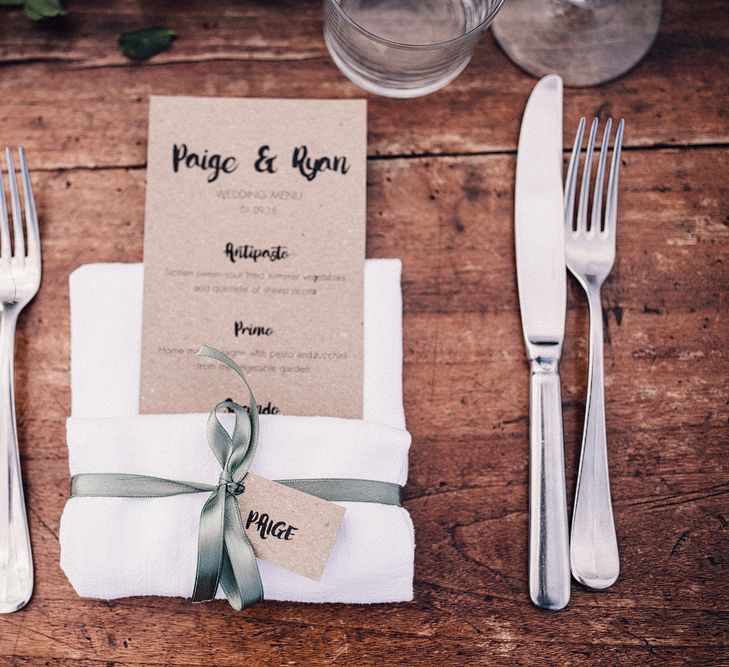 Kraft Paper Wedding Breakfast Menu | Kraft Paper Name Tag | Sage Green Ribbon | Table Runner with Pink Flowers and Foliage | Illustrated Place Setting in White Frame | Rue de Seine Wedding Dress with Pronovias Overskirt, Personalised Place Setting Illustrations and Sweetheart Table | Samuel Docker Photography